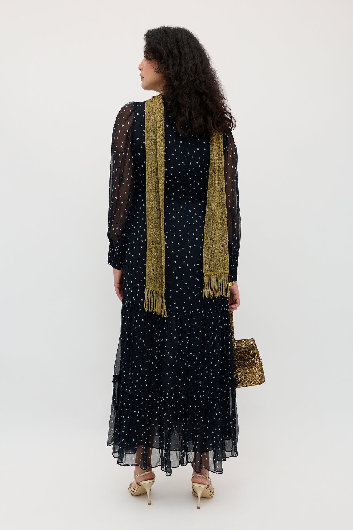 A woman faces away, wearing the Elsie - Starry Night Navy dress with sheer sleeves, gold heels, a gold scarf draped around her neck, and holding a gold handbag against a plain light background.