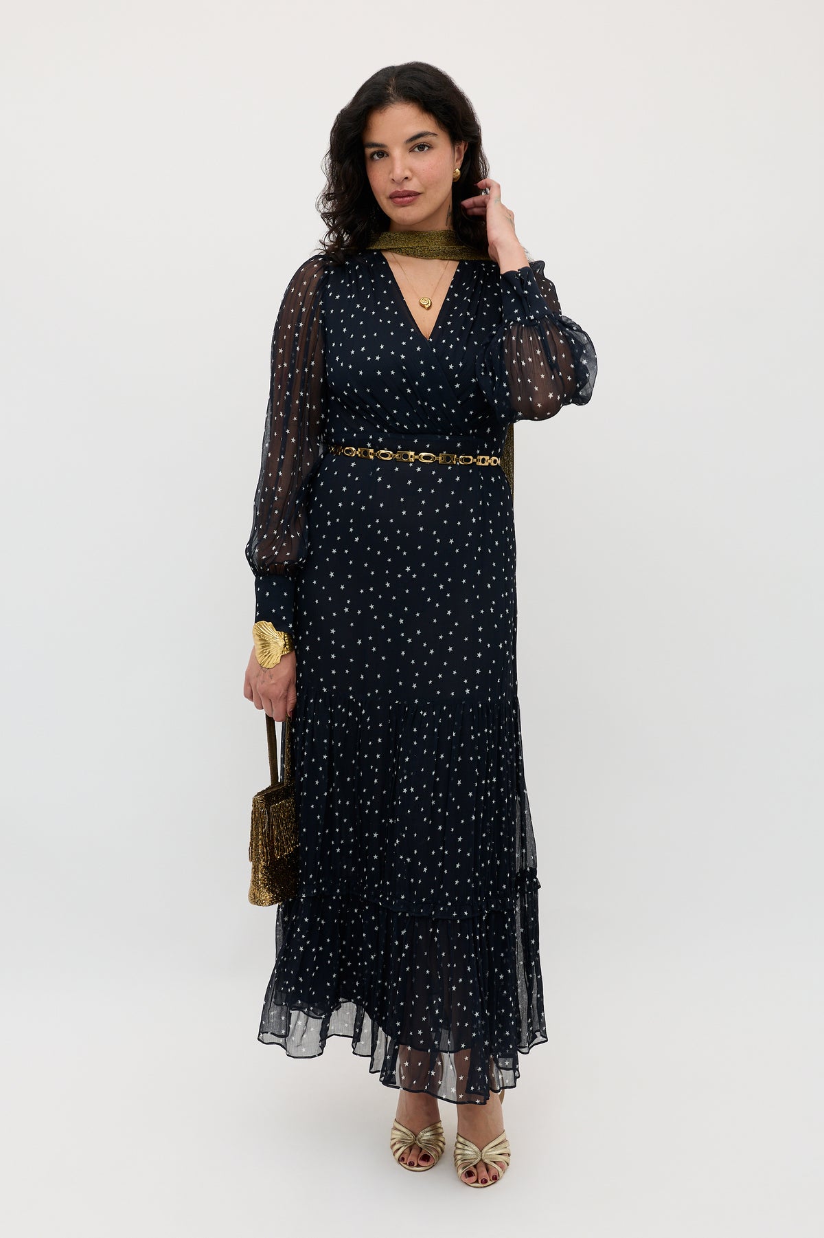A woman stands against a white background wearing the Elsie - Starry Night Navy dress—a long, sheer navy dress with white polka dots—styled with gold jewelry, a chain belt, open-toe heels, and a small gold purse. Her wavy dark hair completes the look.