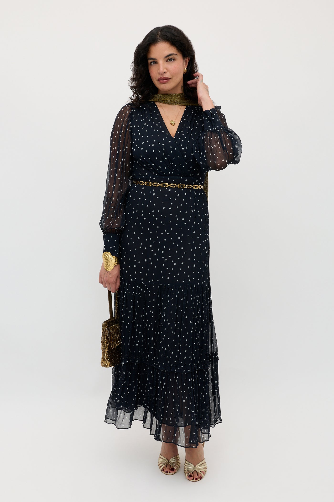 A woman stands against a white background wearing the Elsie - Starry Night Navy dress—a long, sheer navy dress with white polka dots—styled with gold jewelry, a chain belt, open-toe heels, and a small gold purse. Her wavy dark hair completes the look.