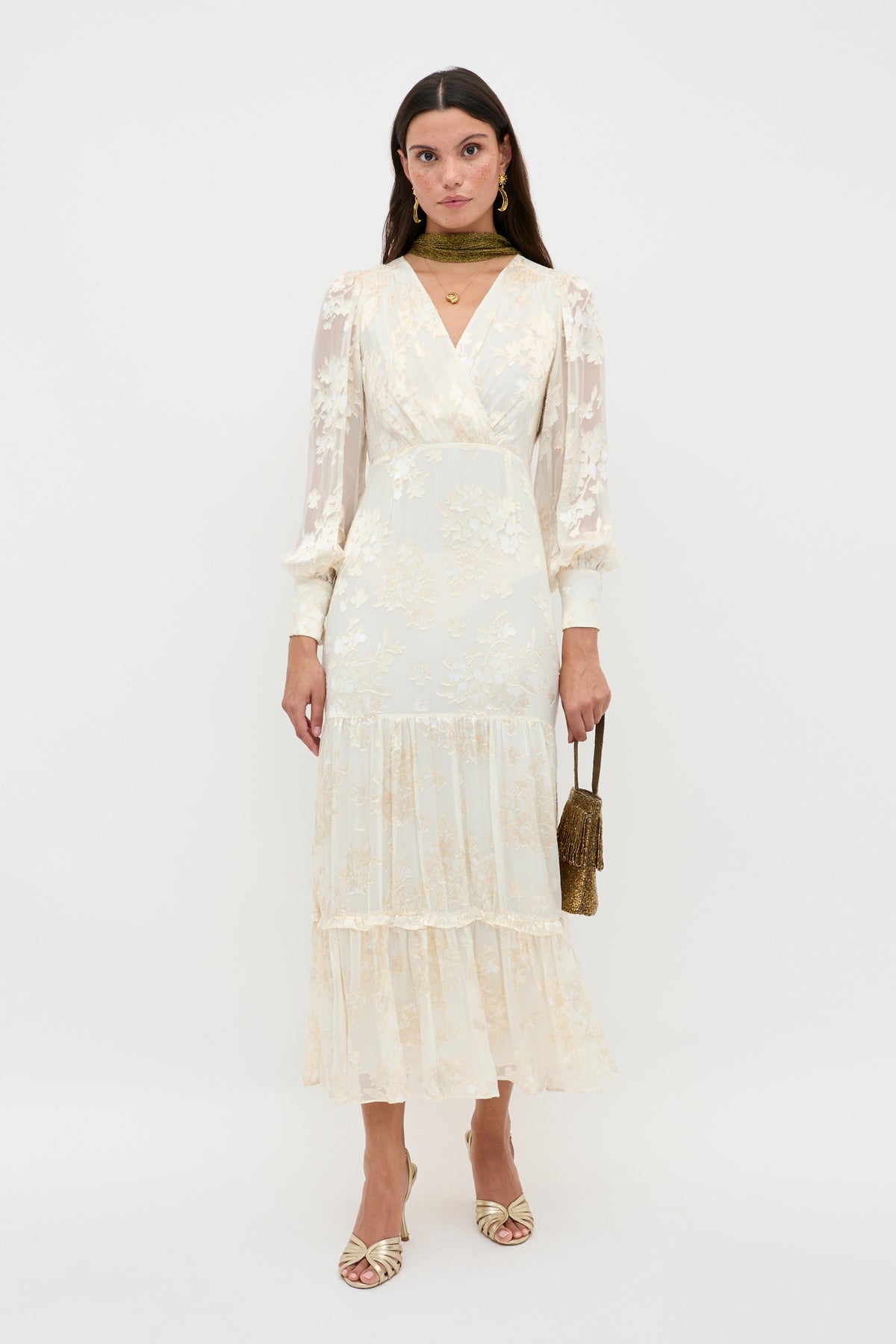 A woman poses against a white background in the Elsie – Bouquet Devore Cream dress, featuring a long, floral-patterned design with sheer sleeves and a tiered skirt, accessorized with gold jewelry, scarf, a small bag, and matching heels.