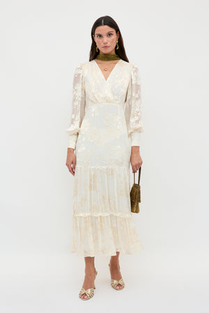 A woman poses against a white background in the Elsie – Bouquet Devore Cream dress, featuring a long, floral-patterned design with sheer sleeves and a tiered skirt, accessorized with gold jewelry, scarf, a small bag, and matching heels.