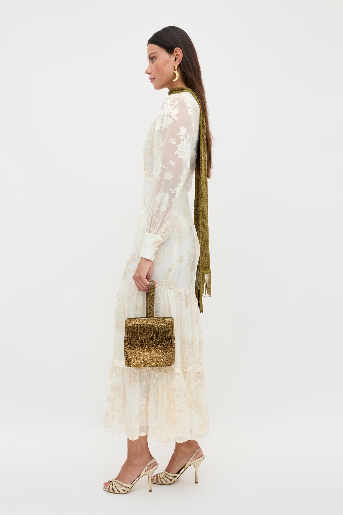 A woman stands in profile against a white background, wearing the Elsie - Bouquet Devore Cream sheer long-sleeve floral midi dress, paired with gold heels, a gold scarf, large gold earrings, and holding a metallic gold handbag.