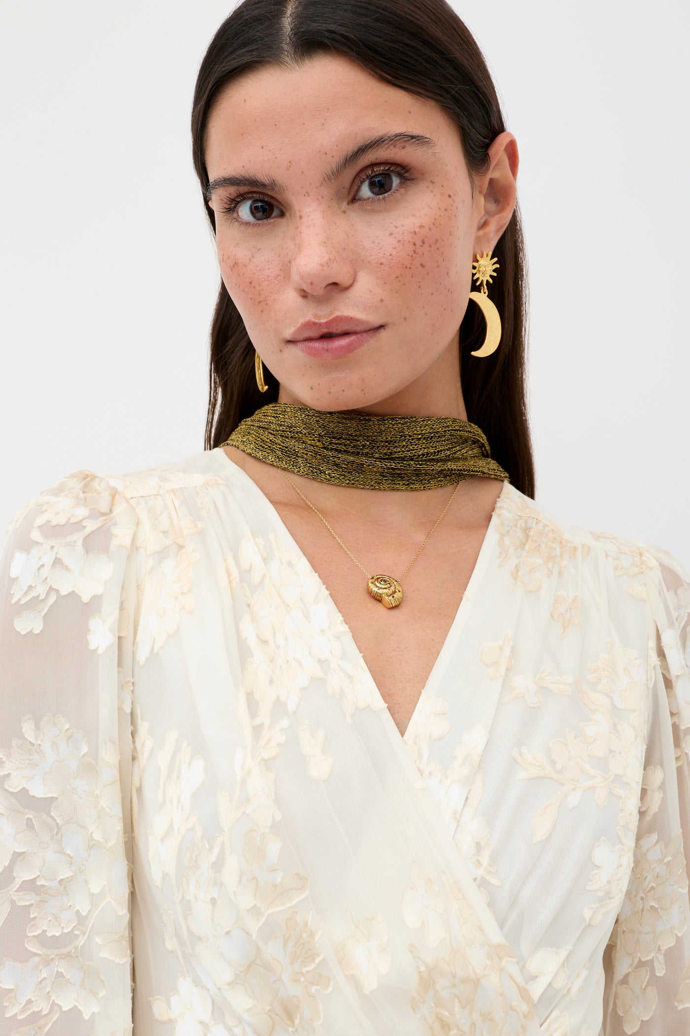 A woman with straight brown hair and freckles wears the Elsie - Bouquet Devore Cream dress, gold crescent moon and sun earrings, a round pendant necklace, and a metallic gold scarf around her neck.