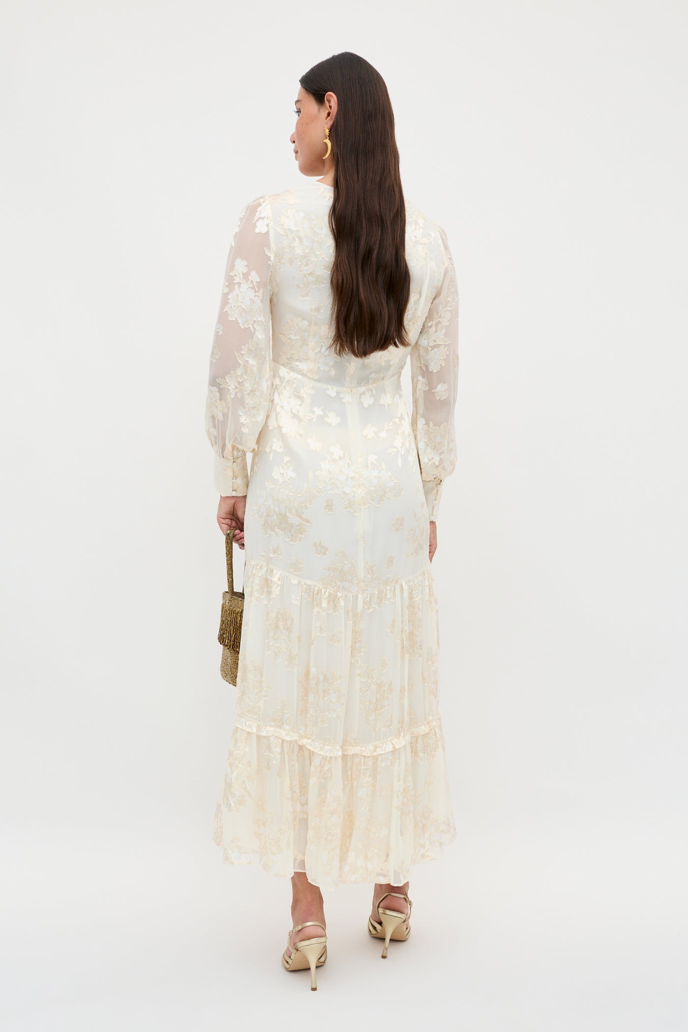 A woman with long brown hair faces away, wearing the Elsie - Bouquet Devore Cream: a sheer, cream floral long-sleeve dress, styled with gold heels and a small gold handbag against a white background.