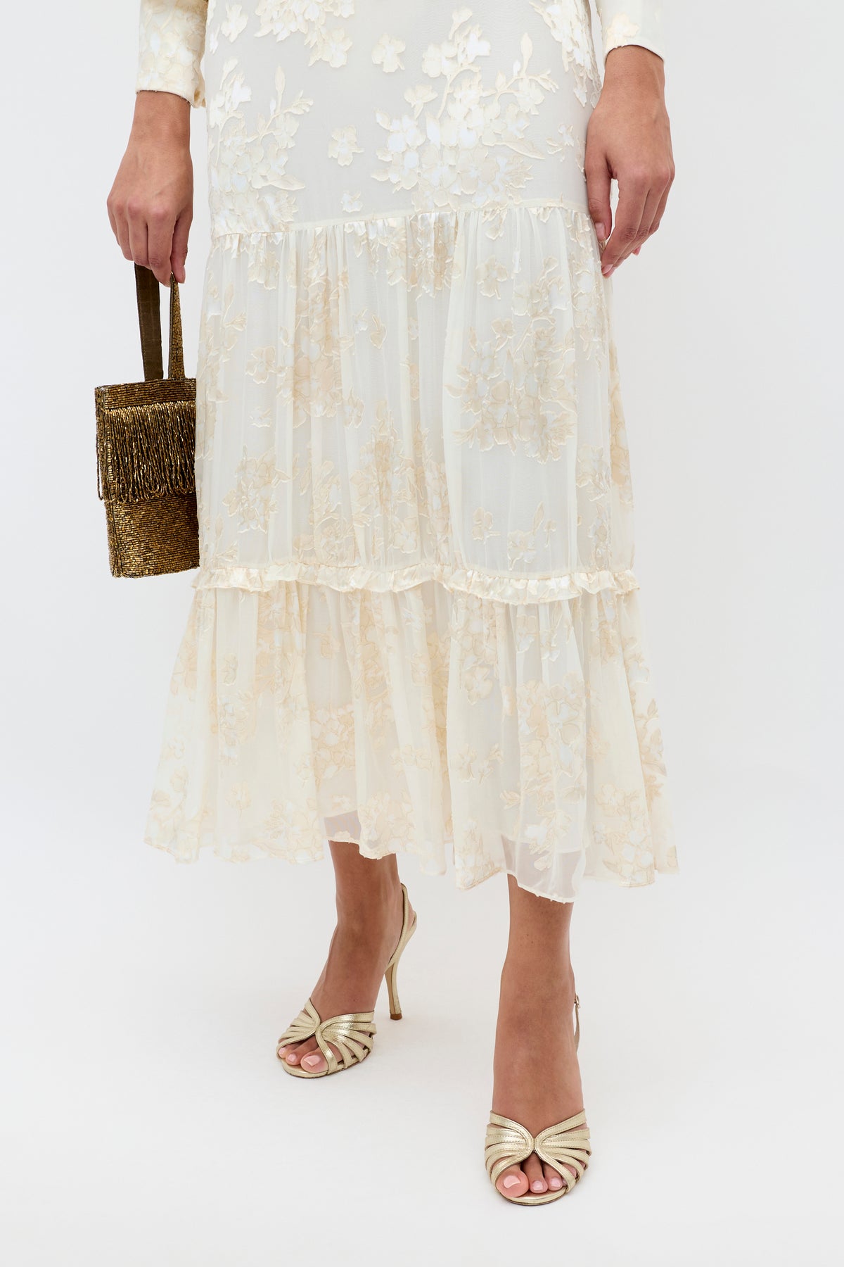 A person wearing the Elsie - Bouquet Devore Cream sheer floral midi dress holds a small gold beaded handbag and wears gold open-toe high-heeled sandals. The image is cropped at the shoulders.