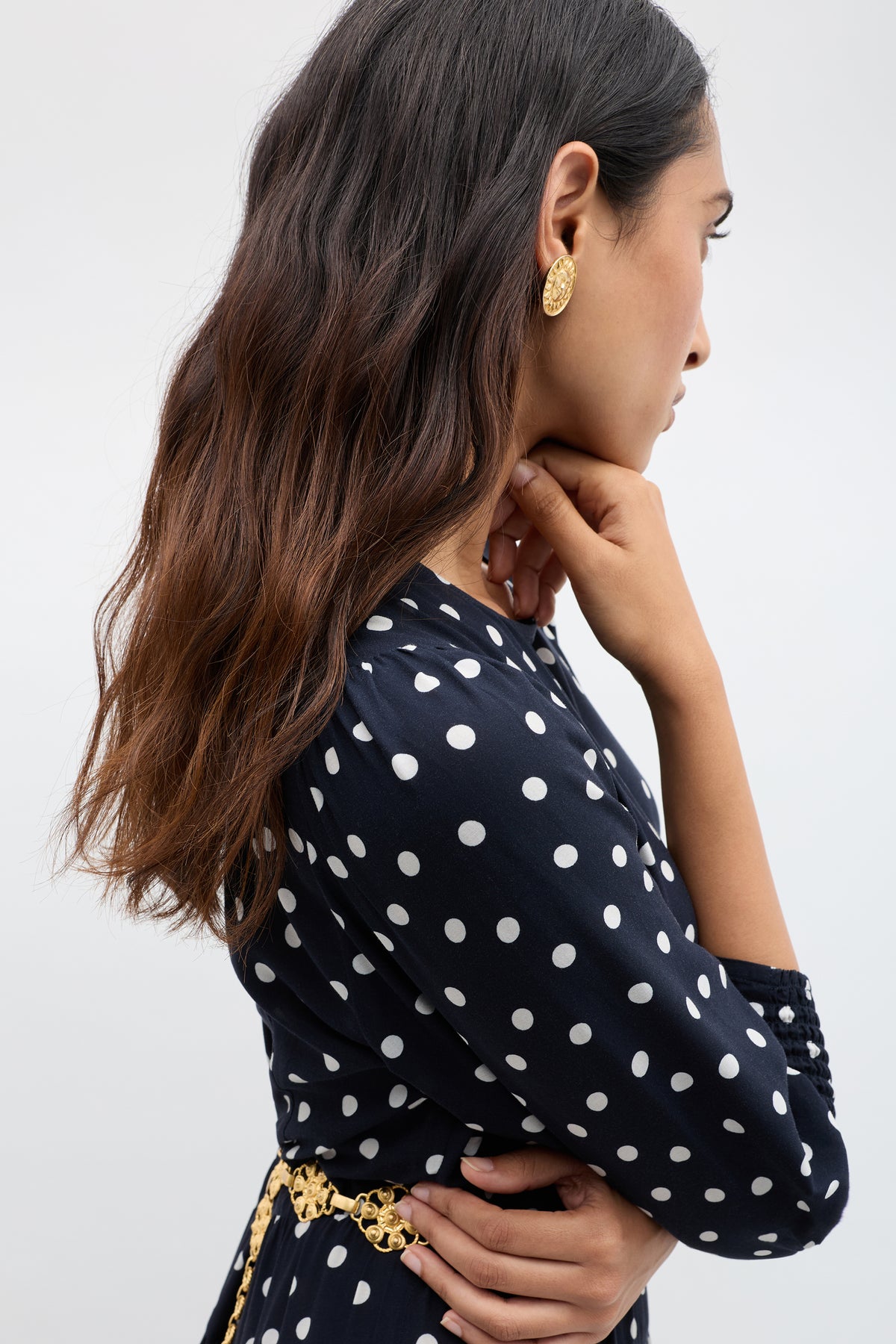 A woman with long, wavy brown hair wears the Kristen - Bohemia Spot Navy dress in profile, featuring a vintage-inspired navy blue fabric with bohemia dots, a gold belt, and gold earrings, posing with her hand near her chin against a plain background.