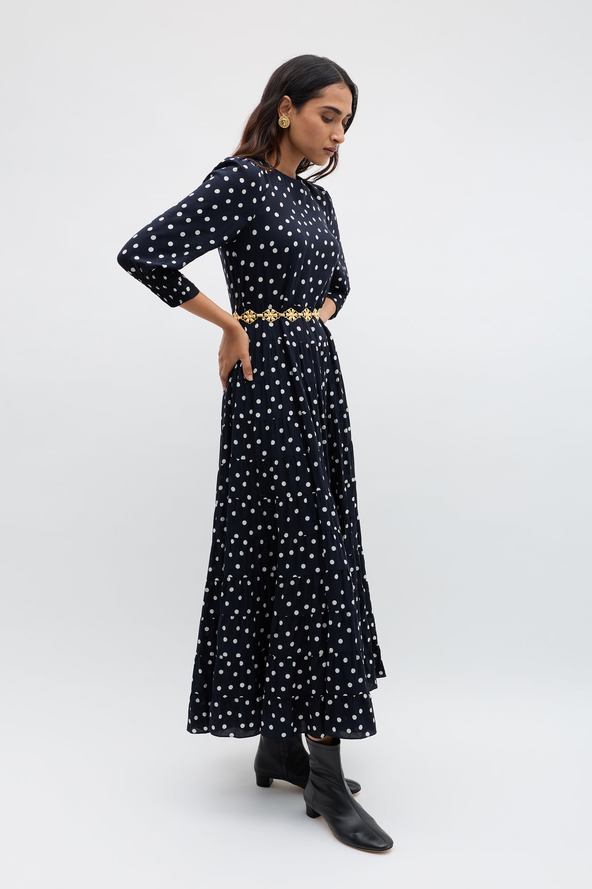 Woman standing in a side pose wearing the Kristen - Bohemia Spot Navy dress, styled with a gold belt, black ankle boots, and large earrings against a plain light backdrop.