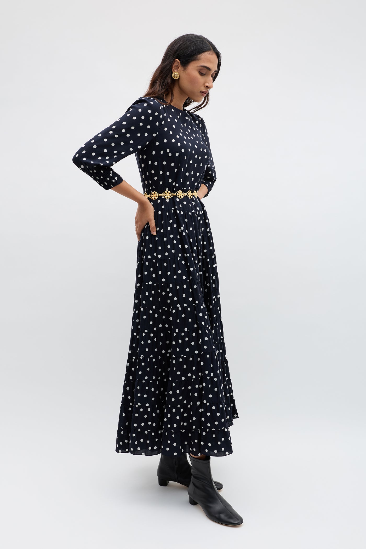 Woman standing in a side pose wearing the Kristen - Bohemia Spot Navy dress, styled with a gold belt, black ankle boots, and large earrings against a plain light backdrop.