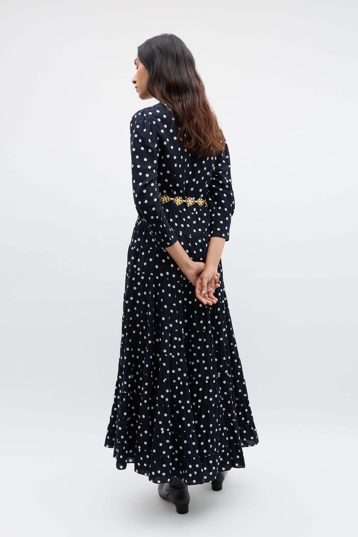 A woman with long brown hair stands facing away, wearing the Kristen - Bohemia Spot Navy dress with a vintage look and bohemian spot print. Her hands are clasped behind her back, and she pairs it with black shoes against a plain background.