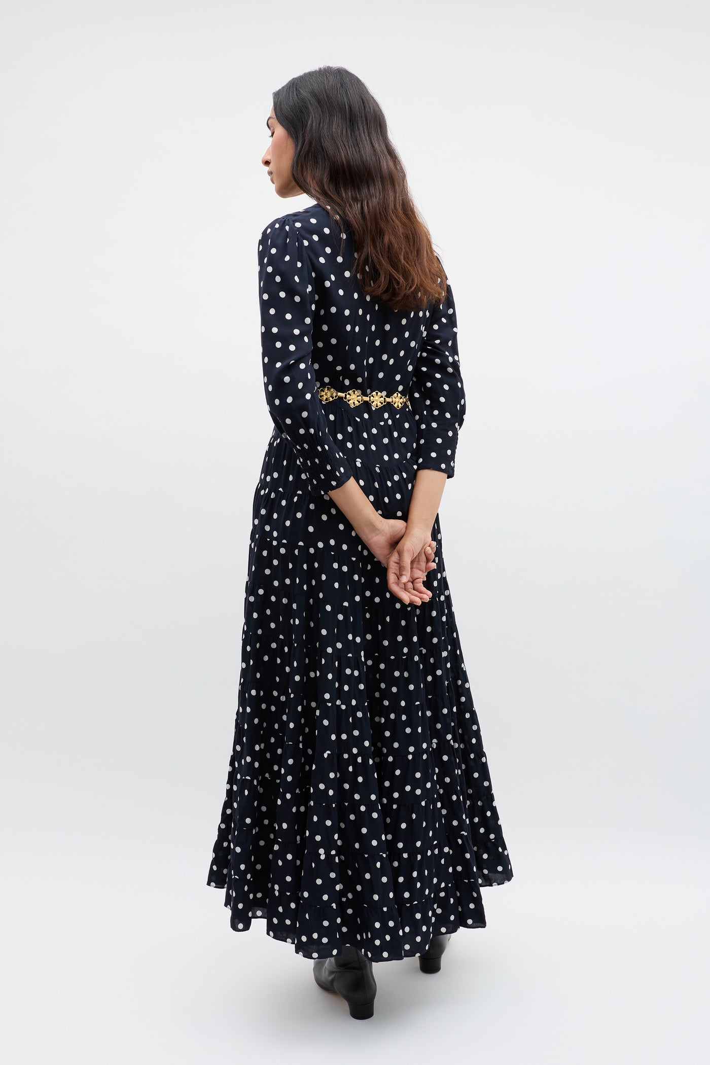 A woman with long brown hair stands facing away, wearing the Kristen - Bohemia Spot Navy dress with a vintage look and bohemian spot print. Her hands are clasped behind her back, and she pairs it with black shoes against a plain background.