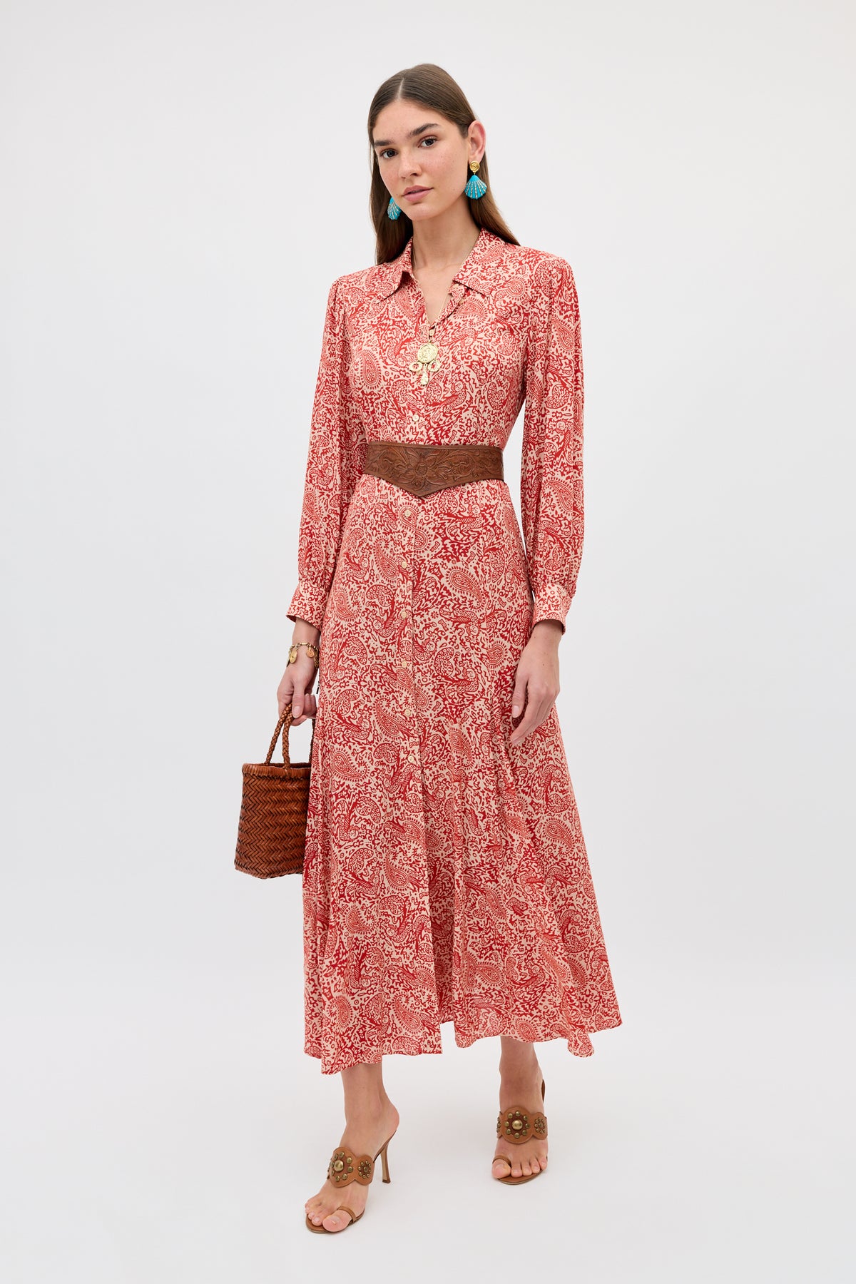 A woman in the Maddison - Woodcut Gremlin Red long-sleeved midi dress, brown belt, tan heeled sandals, turquoise earrings, and a brown woven handbag stands against a plain white background.
