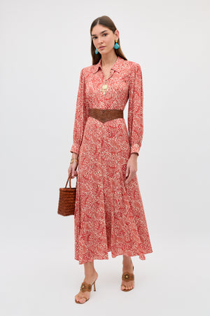 A woman in the Maddison - Woodcut Gremlin Red long-sleeved midi dress, brown belt, tan heeled sandals, turquoise earrings, and a brown woven handbag stands against a plain white background.