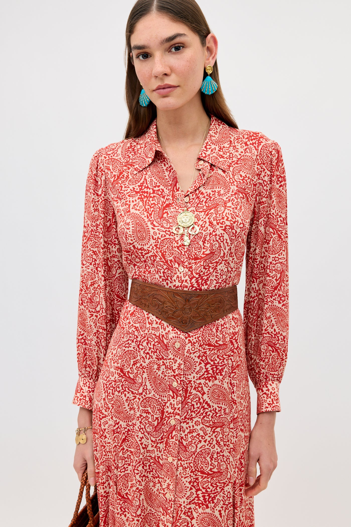 A woman stands against a plain background wearing the Maddison - Woodcut Gremlin Red dress, styled with a brown wide belt, turquoise tasseled earrings, layered gold necklaces, and holding a brown woven bag.
