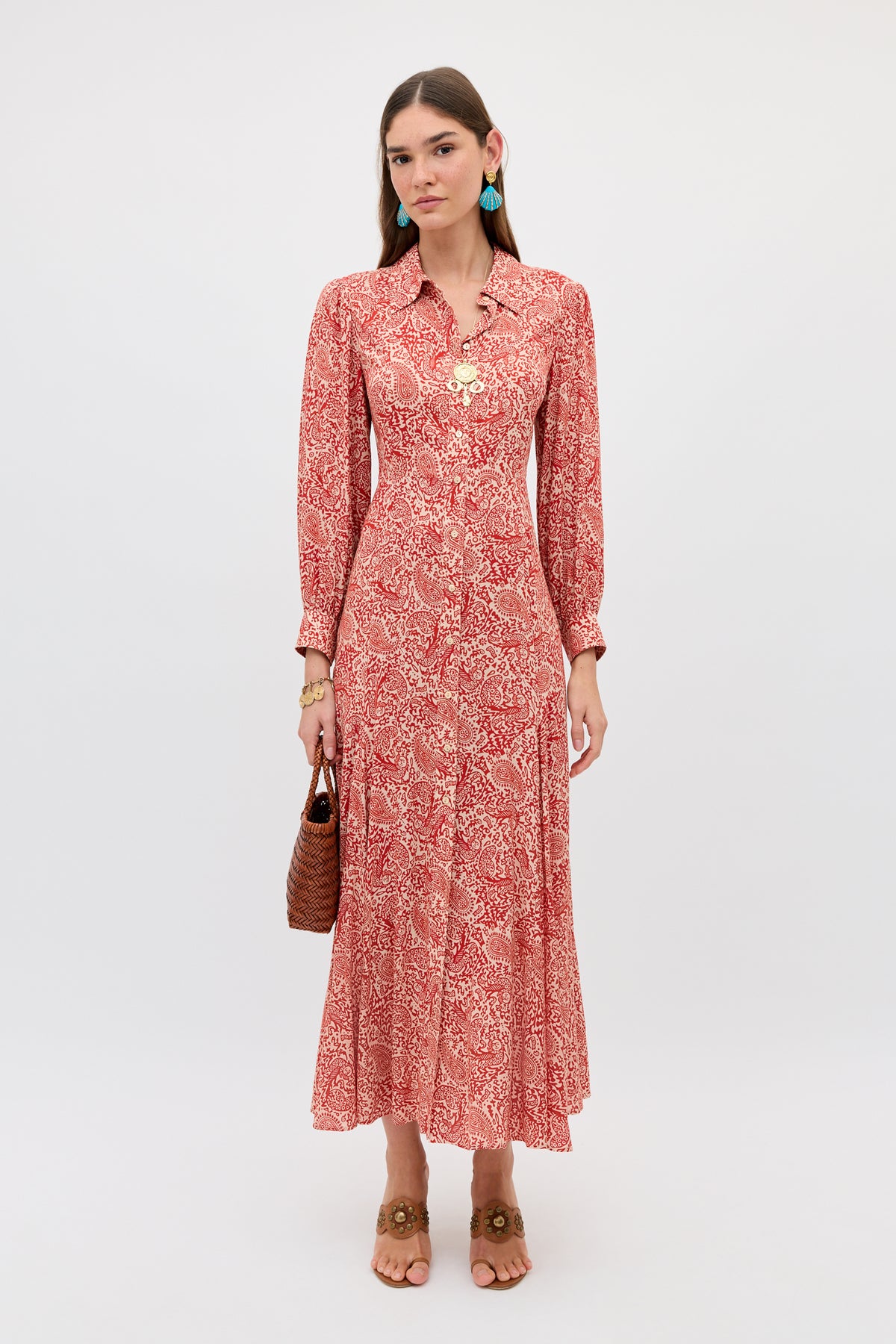 A woman with straight brown hair poses against a white background wearing the Maddison - Woodcut Gremlin Red long-sleeved maxi dress, styled with a woven brown handbag, tan sandals, blue earrings, and gold jewelry.