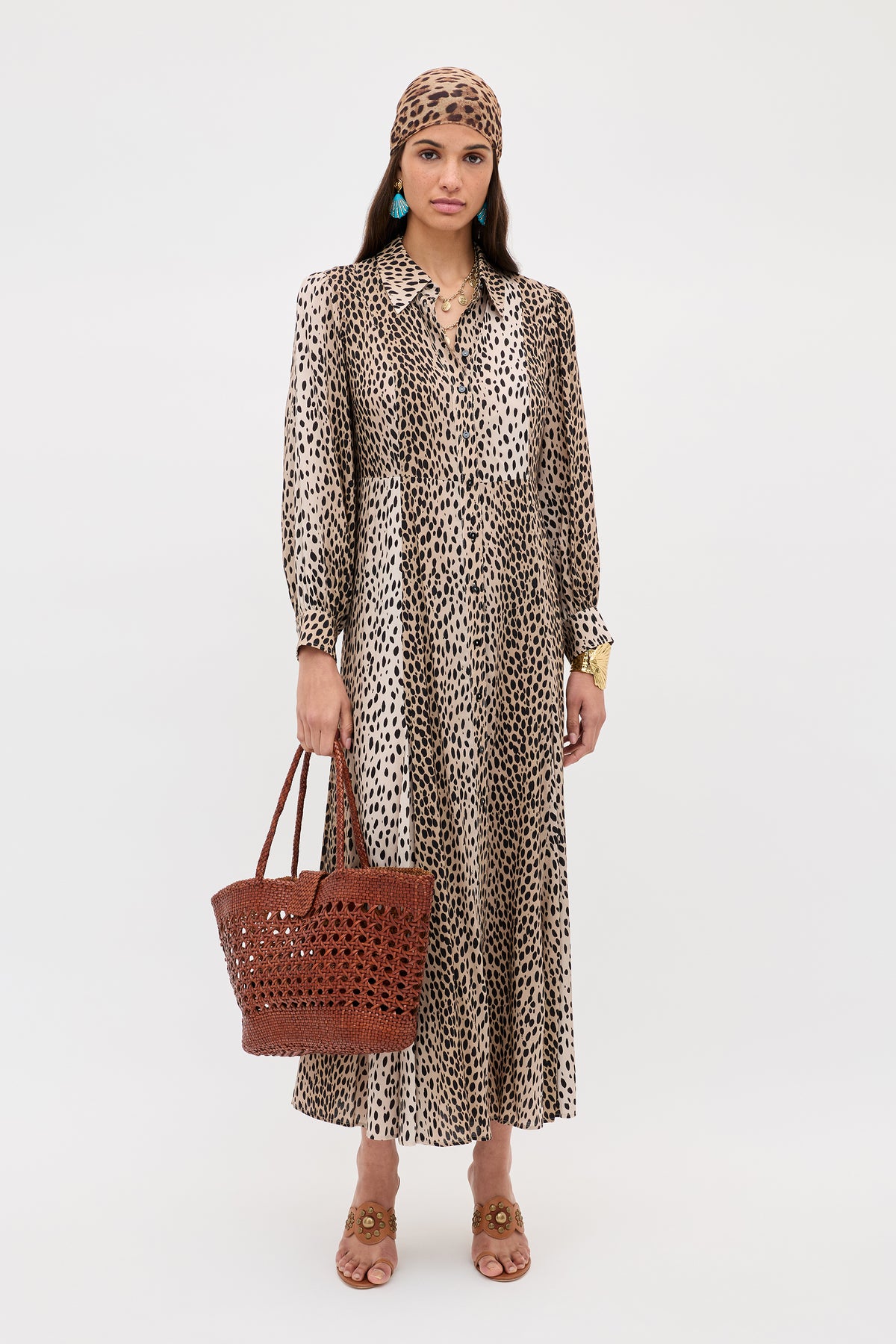A petite woman stands against a white background wearing the Maddison - Antelope Spot Black long, loose animal-print shirt dress with a matching headscarf, tan sandals, turquoise earrings, and a brown woven tote bag—ideal for everyday style.