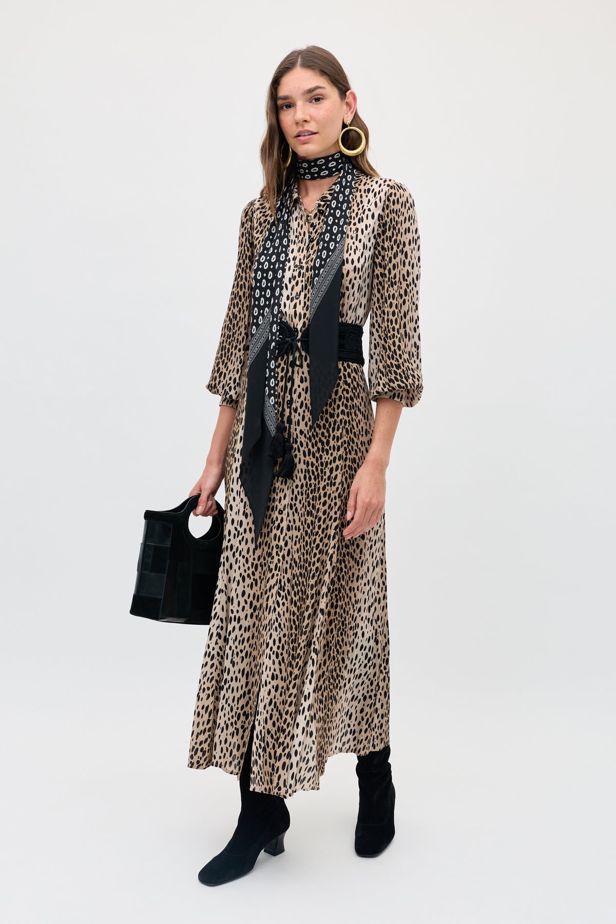 A woman stands against a plain white background wearing the Maddison - Antelope Spot Black, a long, leopard-print shirt dress perfect for everyday style. She pairs it with black boots, a patterned scarf, gold hoop earrings, and carries a black handbag. This look is available in both regular and petite sizes.