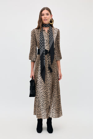 A woman stands against a plain white background wearing the Maddison - Antelope Spot Black long shirt dress, perfect for both regular and petite everyday styles, paired with black boots, a patterned scarf, large hoop earrings, and holding a black handbag.