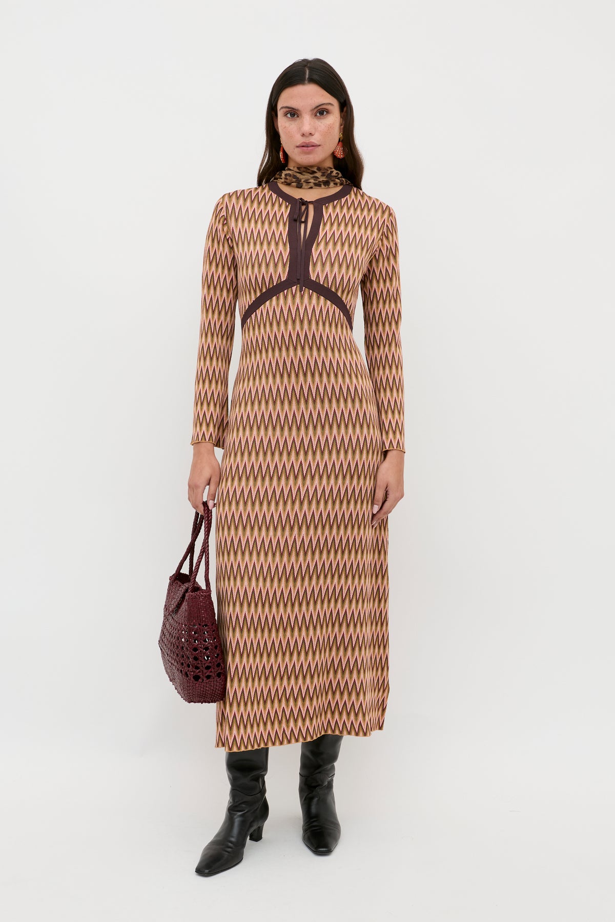 A woman stands against a white background wearing the Pamela - Maeve Jacquard Multi dress, which features long sleeves and an ankle-length geometric zigzag pattern in beige and brown, paired with black boots and a dark red handbag.