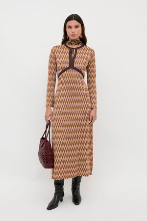 A woman stands against a white background wearing the Pamela - Maeve Jacquard Multi dress, which features long sleeves and an ankle-length geometric zigzag pattern in beige and brown, paired with black boots and a dark red handbag.