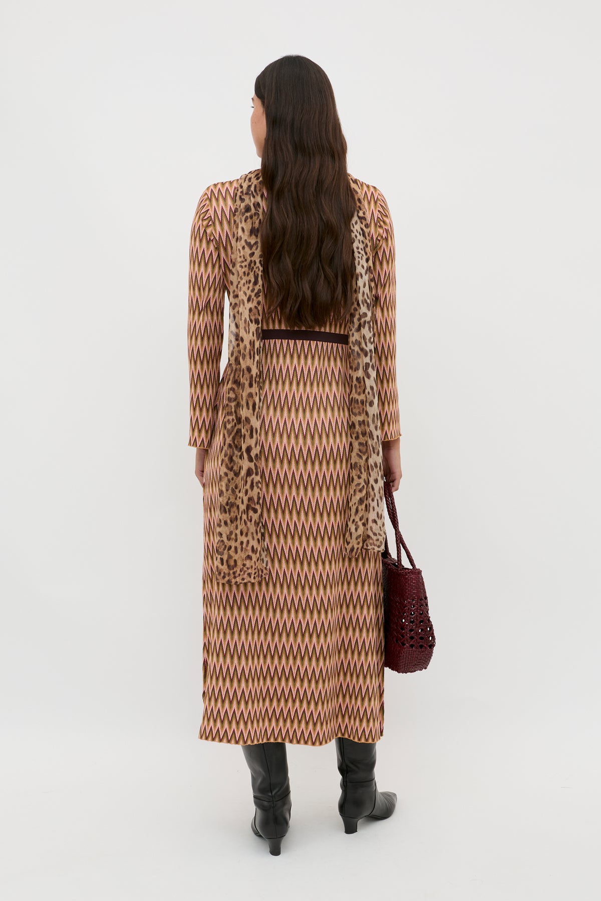 A woman with long, loose dark hair stands facing away on a white background, wearing the Pamela - Maeve Jacquard Multi zigzag-patterned long dress, a leopard scarf, black knee-high boots, and holding a maroon woven handbag.