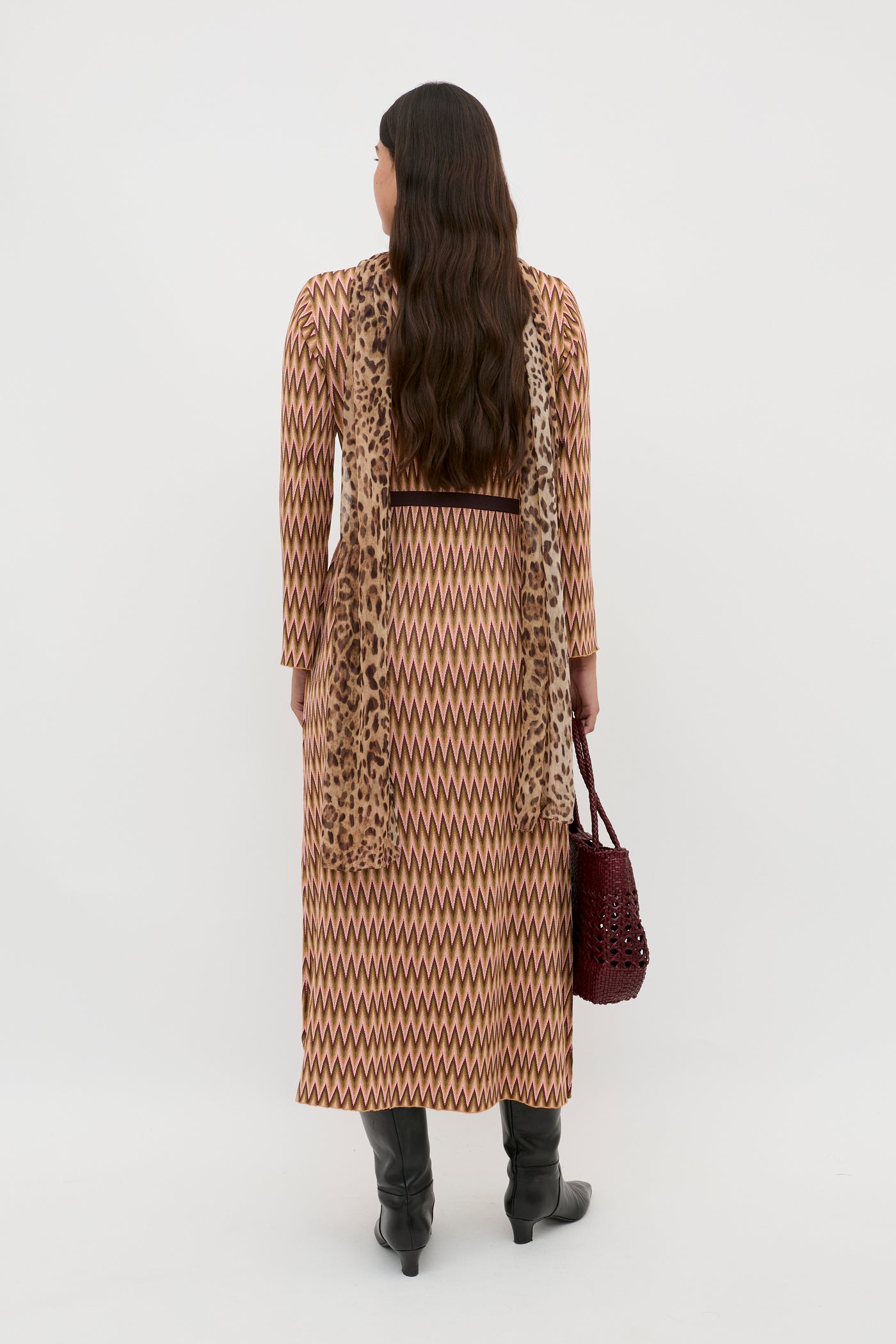 A woman with long, loose dark hair stands facing away on a white background, wearing the Pamela - Maeve Jacquard Multi zigzag-patterned long dress, a leopard scarf, black knee-high boots, and holding a maroon woven handbag.
