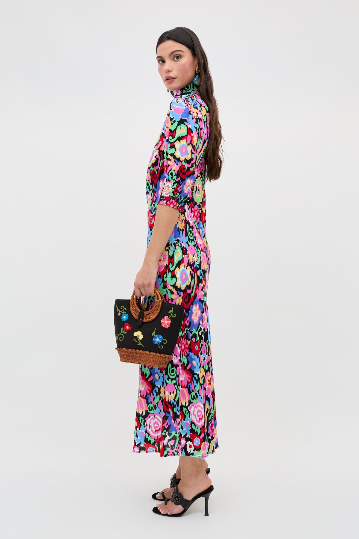 A woman stands sideways in the Jess - Ikat Bloom Charcoal dress with elbow-length sleeves, holding a black round-handle handbag and wearing black heeled sandals against a plain white background.