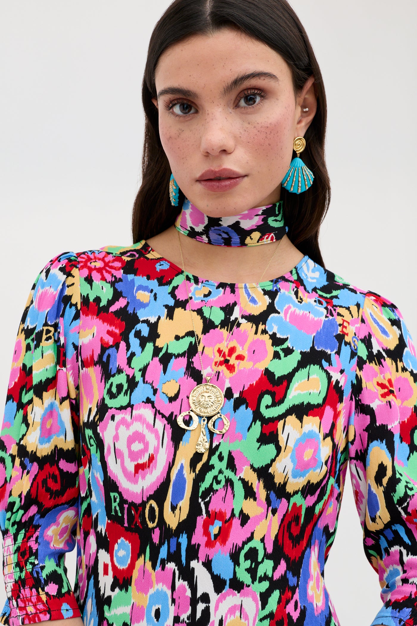 A woman wearing the "Jess - Ikat Bloom Charcoal" dress, paired with a matching neck scarf, blue tassel earrings, and a round gold pendant necklace stands against a plain light background.