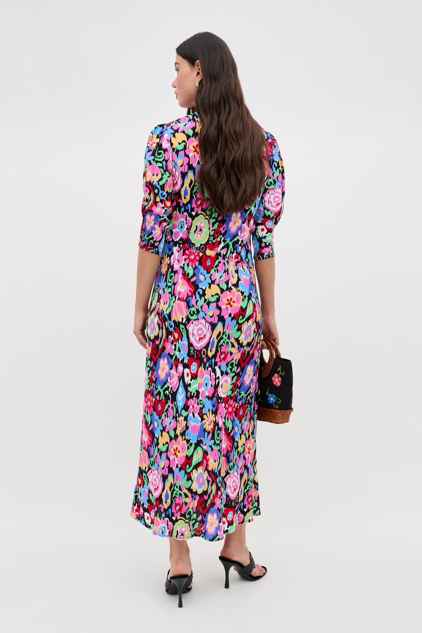 A woman with long brown hair faces away, wearing the Jess - Ikat Bloom Charcoal dress with black heeled sandals and a small black handbag. The background is plain and light.