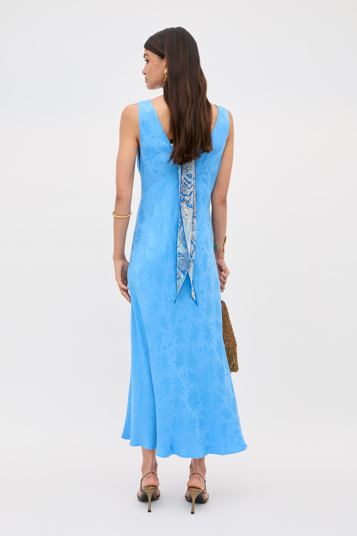 A woman with long brown hair and a patterned scarf wears the Sandrine - Daisy Jacquard Blue V-neck midi slip dress by RIXO, paired with high-heeled sandals and a brown handbag.