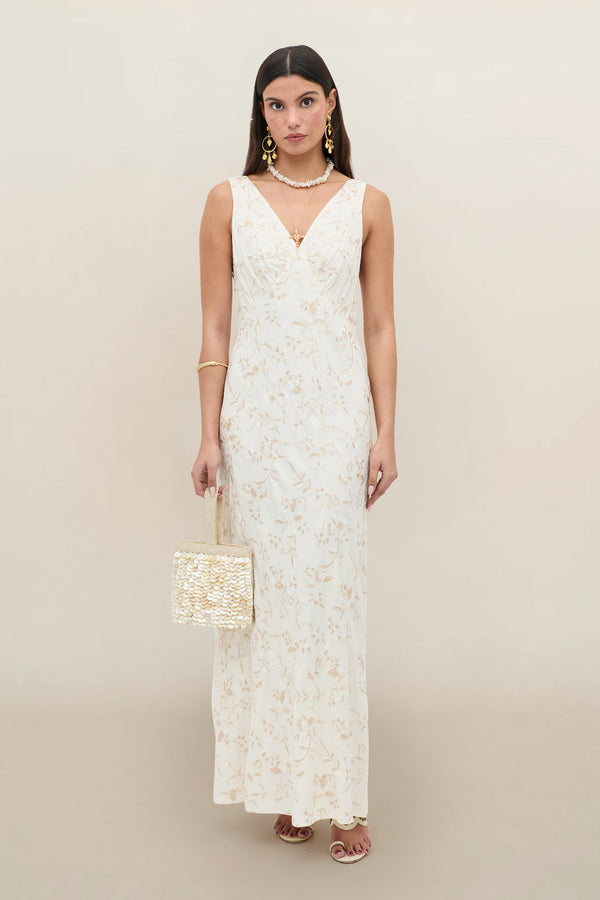 A woman wears the Fawn - Botanical Embroidery Pearl sleeveless, floor-length white dress with subtle floral patterns, styled with a woven handbag, gold jewelry, and heeled sandals against a light background.