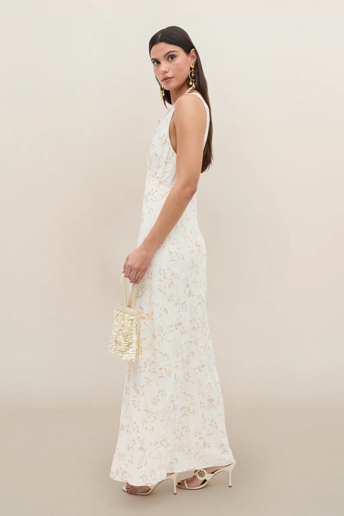 A woman wears the Fawn - Botanical Embroidery Pearl: a sleeveless, floor-length, light cream form-fitting dress with subtle floral embroidery; she stands sideways, accessorized with gold earrings, white heels, and a small woven handbag.