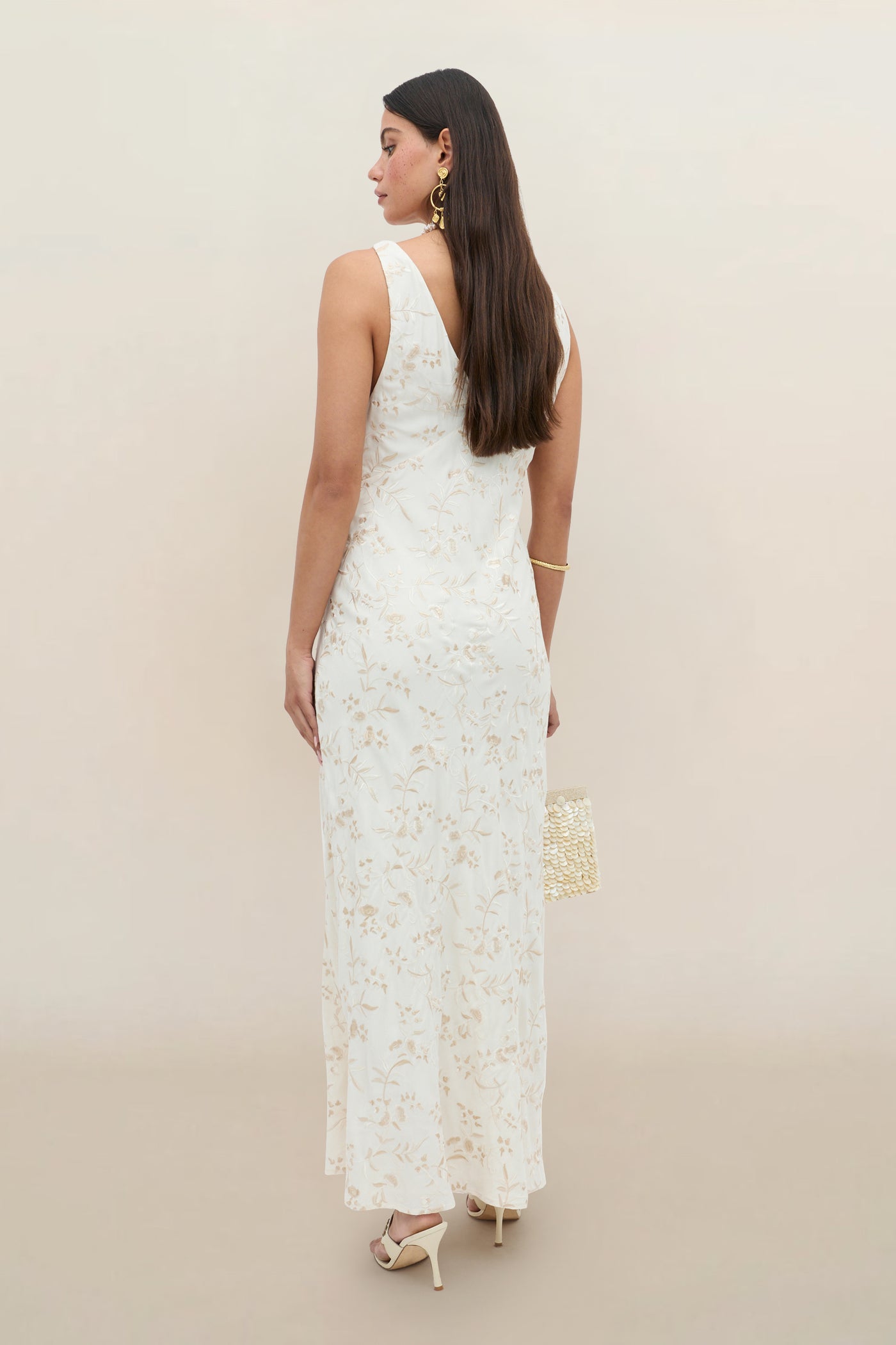 Wearing the Fawn - Botanical Embroidery Pearl, a sleeveless, ankle-length white dress with floral embroidery, she stands with her back to the camera, holding a beaded bag and accessorizing with heeled sandals and gold earrings.