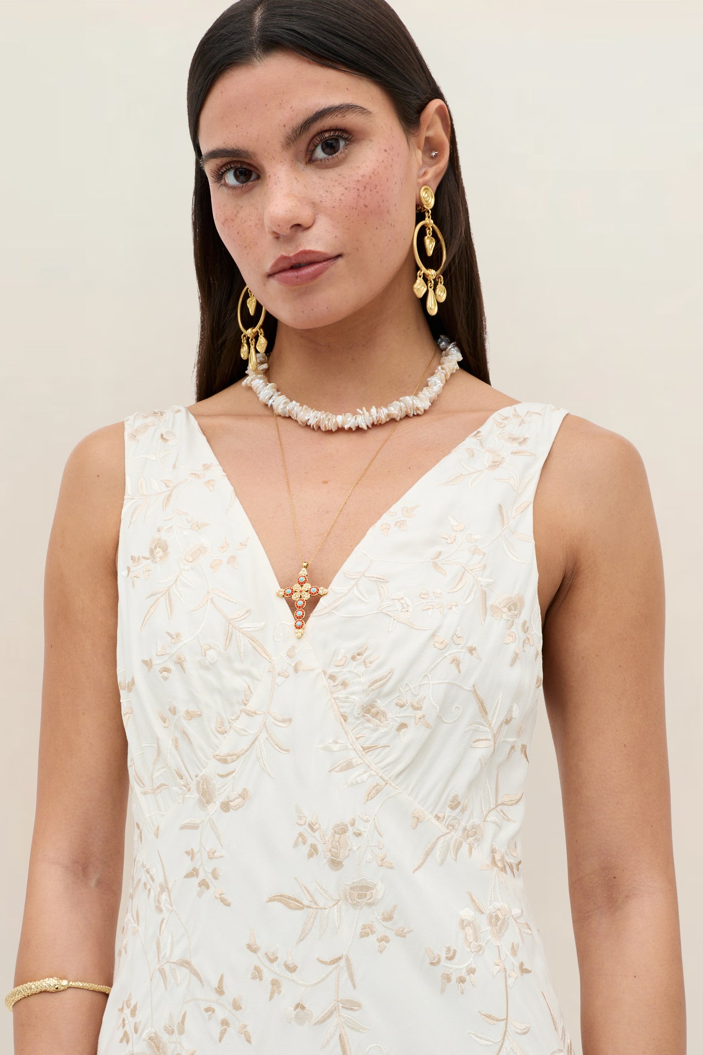 A woman with long dark hair wears the Fawn - Botanical Embroidery Pearl bridal dress, featuring cream fabric and floral embroidery, paired with gold drop earrings, shell and pendant necklaces, and a gold bracelet against a neutral background.