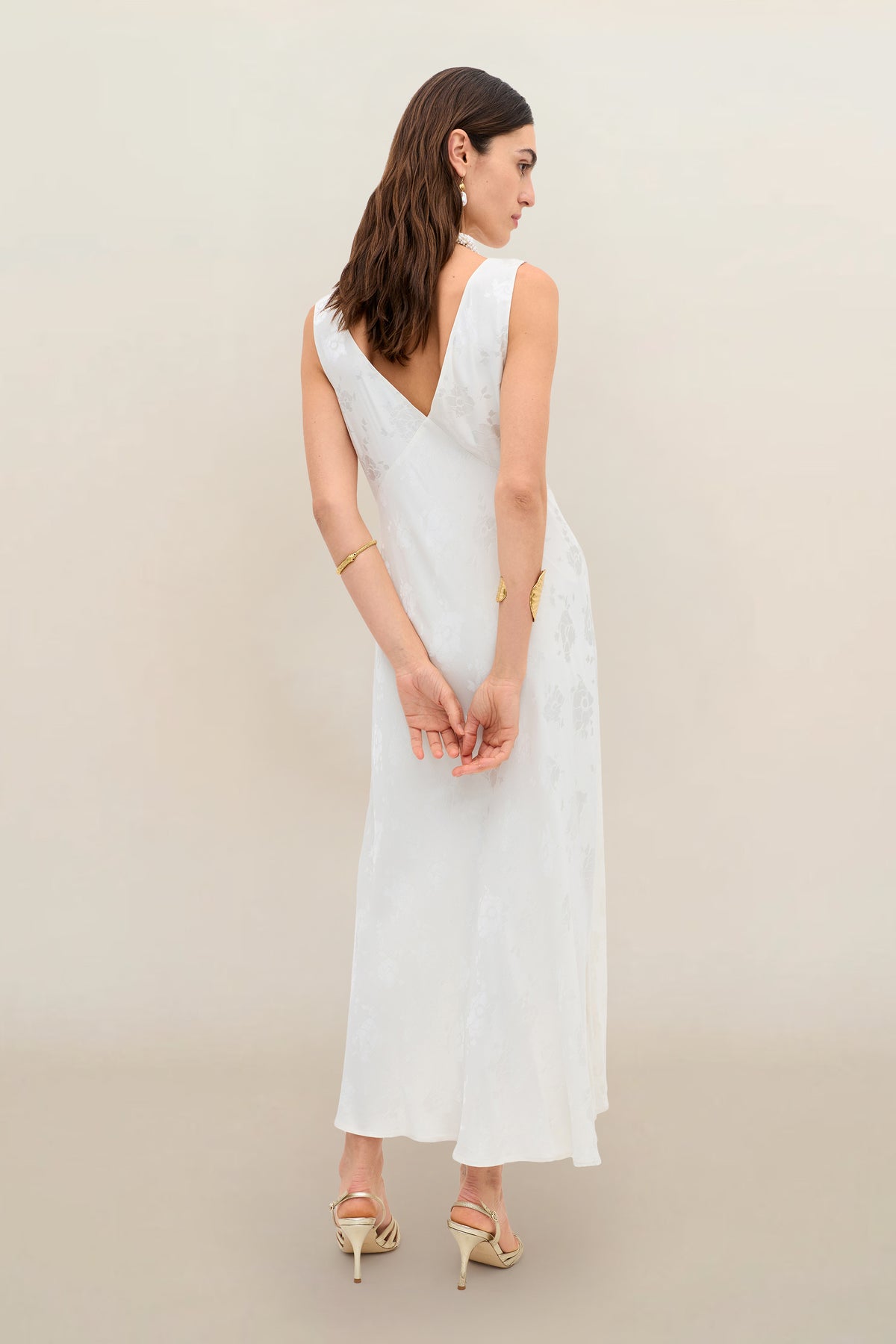 A woman stands with her back to the camera, hands clasped behind her, displaying the Sandrine - Poppy: a sleeveless, vintage-inspired ivory jacquard dress against a plain beige background.