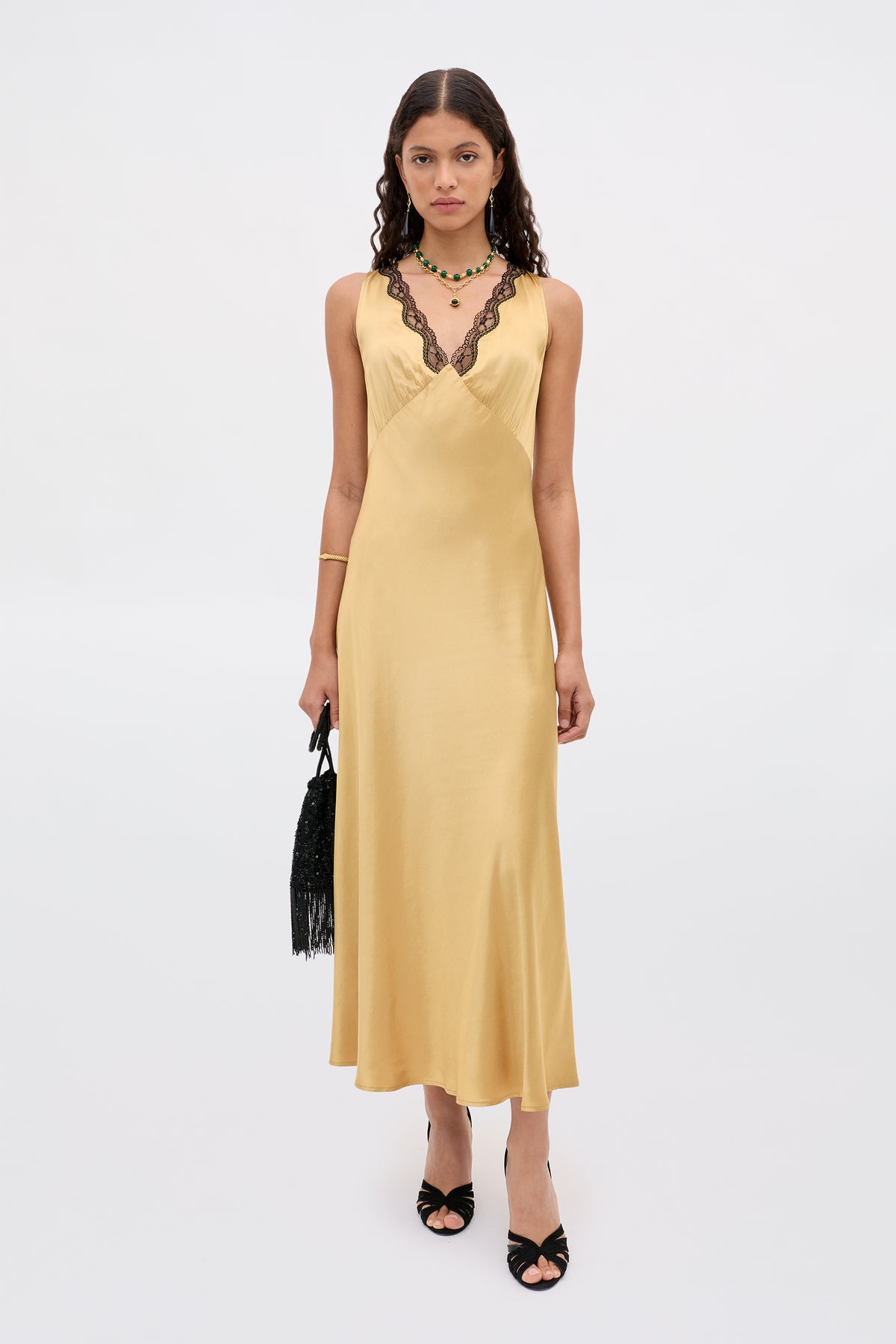 A woman in the Sandrine - Gold long, sleeveless yellow satin dress with black lace trim, paired with black high-heeled sandals, a fringed black handbag, and layered necklaces stands against a plain white background.