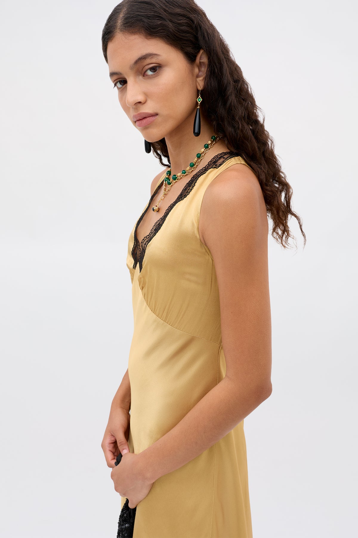 Woman with long curly hair wears the Sandrine - Gold sleeveless yellow satin dress with black lace trim, layered necklaces, and drop earrings. She stands against a plain light background, looking towards the camera.