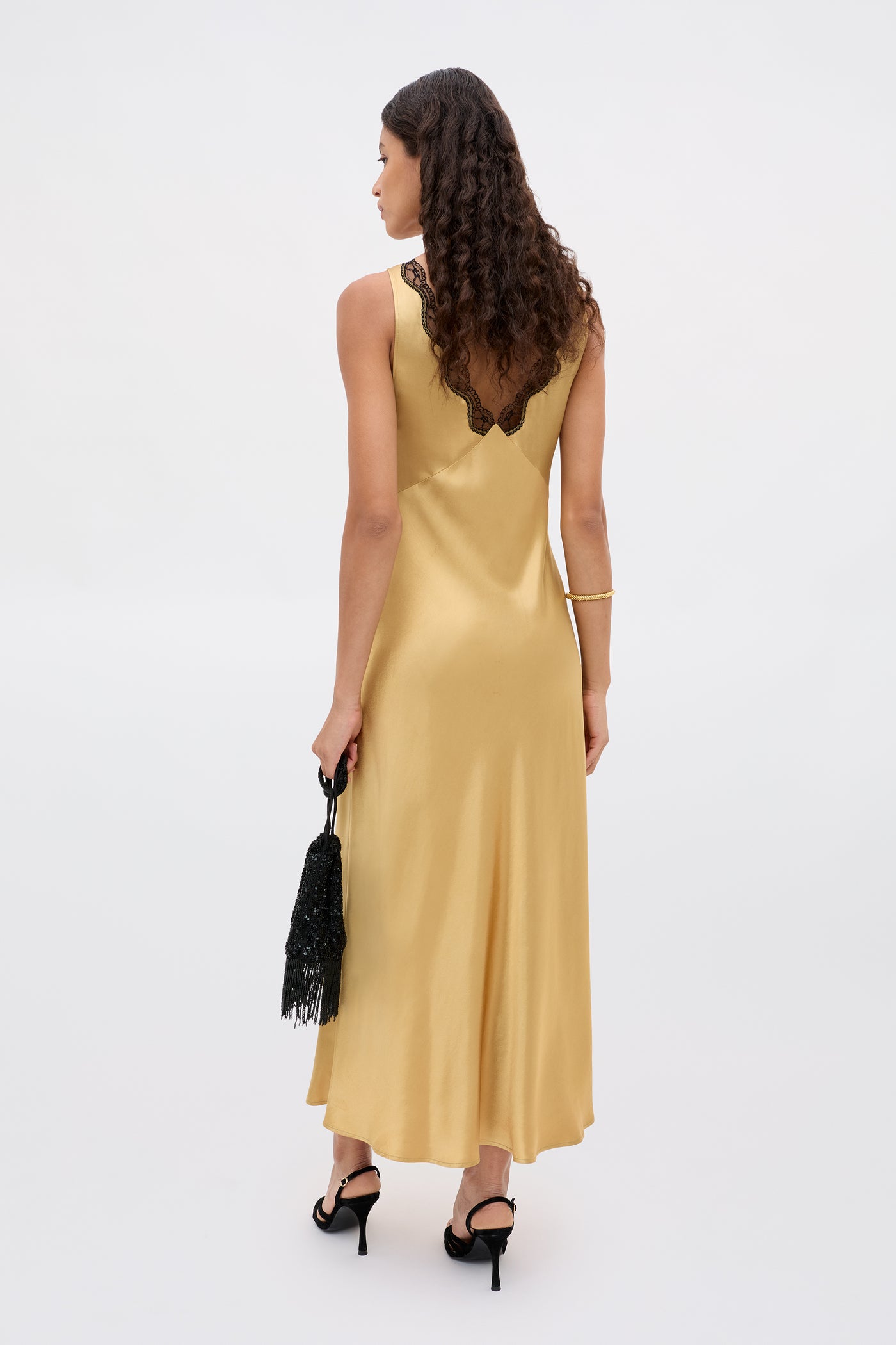 A woman with long curly hair wears the Sandrine - Gold satin sleeveless dress with a deep V-shaped lace back, black heels, and holds a black fringed handbag, standing against a plain light background.