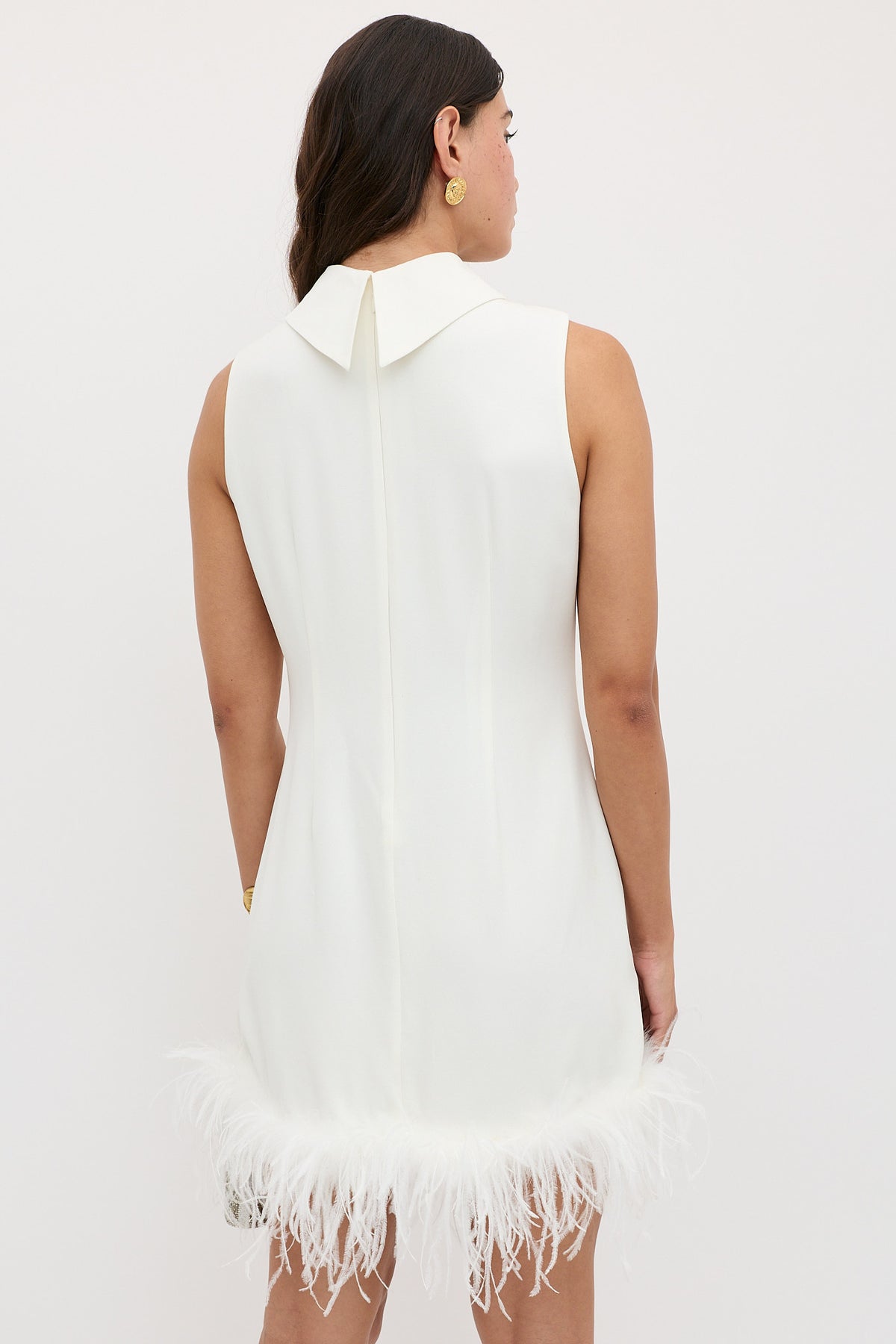 A woman is seen from behind wearing the Candice - Satin Back Silk Crepe dress, a sixties-inspired white shift with a pointed collar and ostrich-feathered hem. She has dark wavy hair, gold earrings, and stands against a plain light background.
