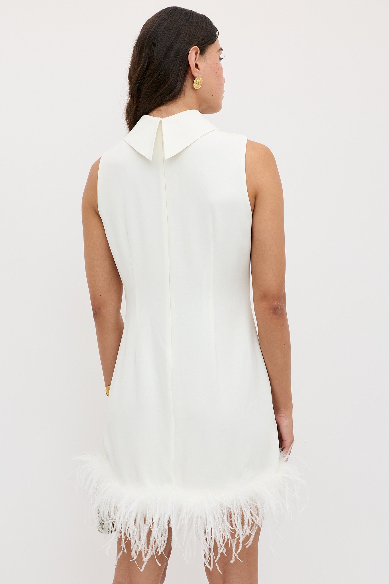 A woman is seen from behind wearing the Candice - Satin Back Silk Crepe dress, a sixties-inspired white shift with a pointed collar and ostrich-feathered hem. She has dark wavy hair, gold earrings, and stands against a plain light background.