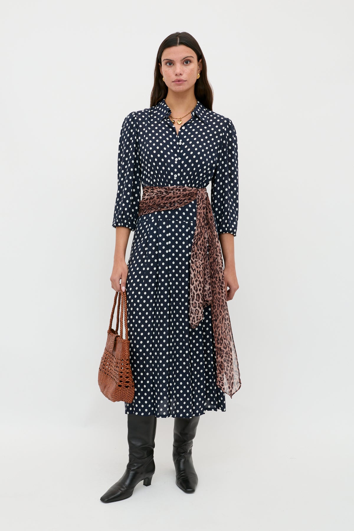 A woman stands against a white background wearing the Izzy - Runway Spot Navy dress, styled with a leopard print sash, black knee-high boots, and holding a brown woven handbag.