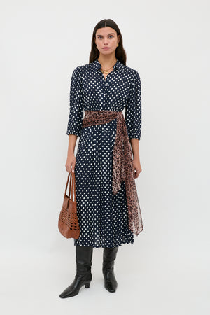 A woman stands against a white background wearing the Izzy - Runway Spot Navy dress, styled with a leopard print sash, black knee-high boots, and holding a brown woven handbag.