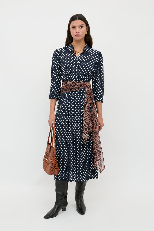 A woman stands against a white background wearing the Izzy - Runway Spot Navy dress, styled with a leopard print sash, black knee-high boots, and holding a brown woven handbag.