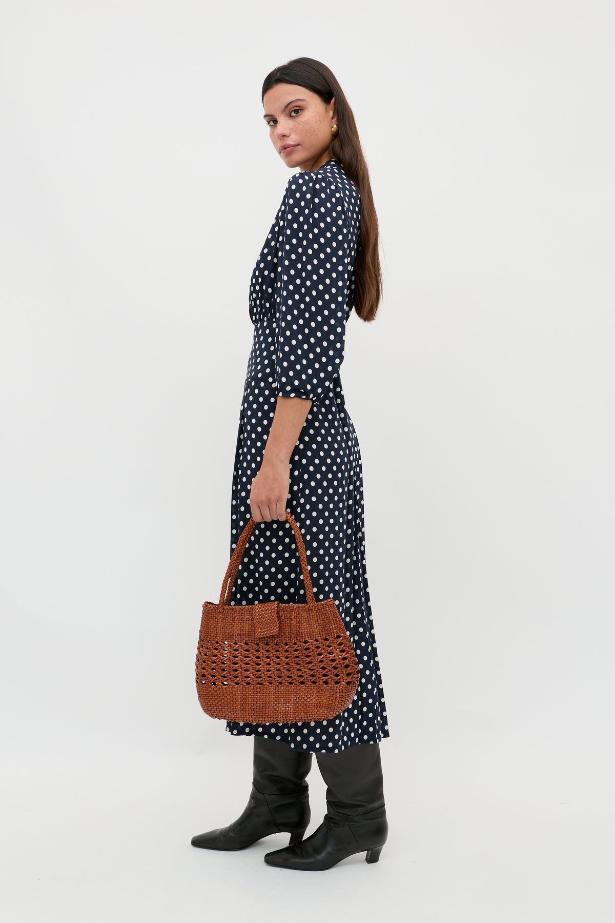 A woman with long dark hair stands sideways against a plain white background, holding a brown woven handbag and wearing the Izzy - Runway Spot Navy shirtdress with blouson sleeves and black knee-high boots.