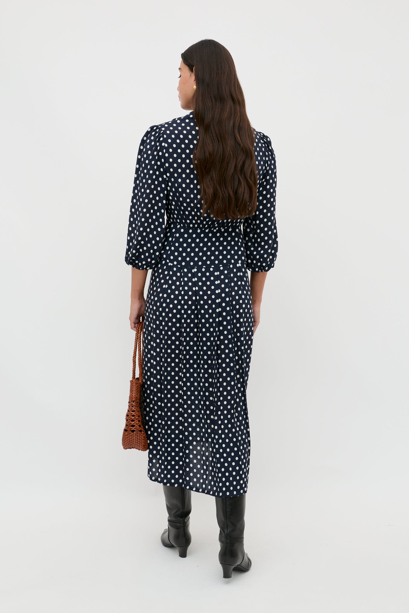 A woman with long brown hair, wearing the Izzy - Runway Spot Navy shirtdress and black boots, stands facing away and holds a brown woven handbag against a plain white background.