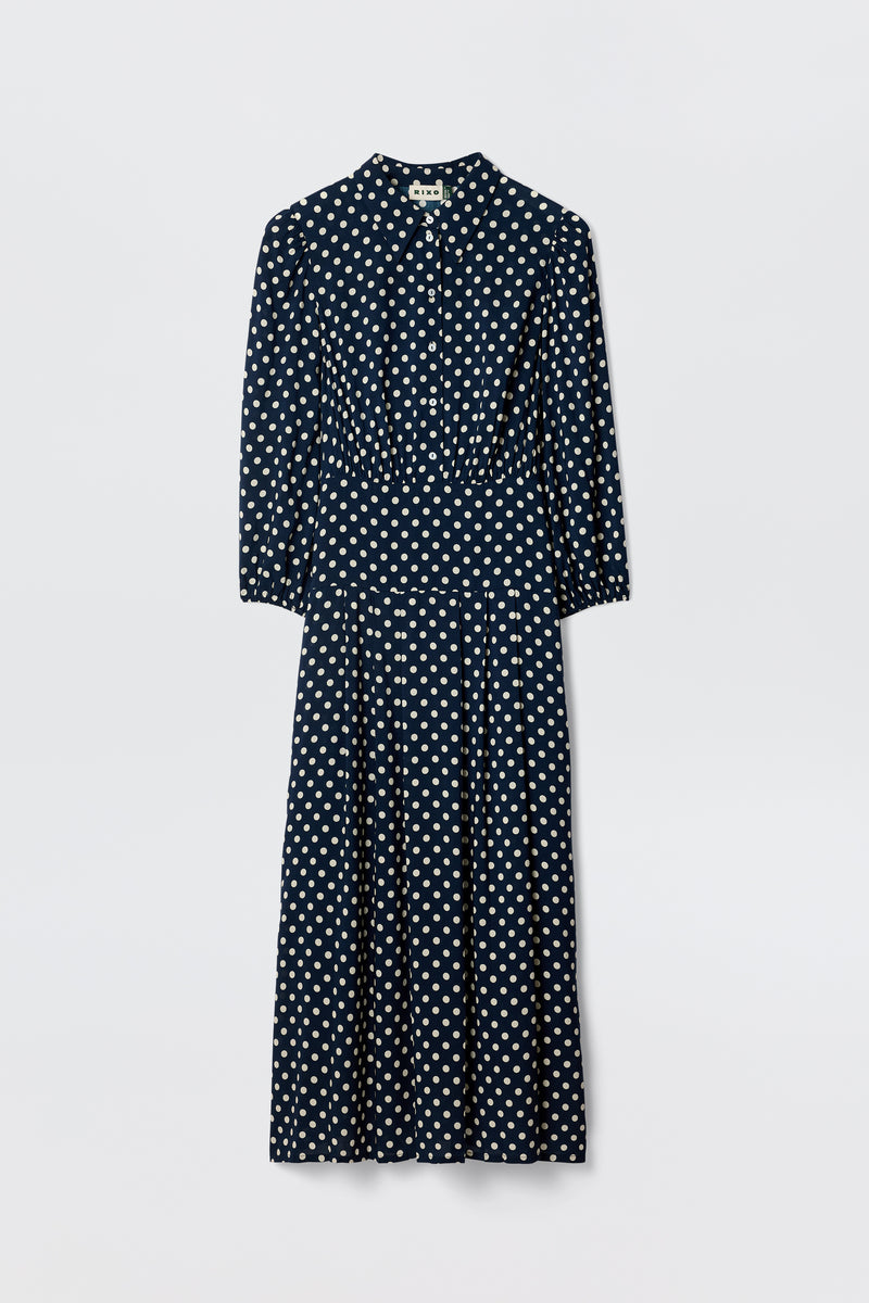 The Izzy - Runway Spot Navy is a navy polka dot shirtdress with long blouson sleeves, a collared button-down front, gathered waist, and midi skirt, shown on a plain white background.