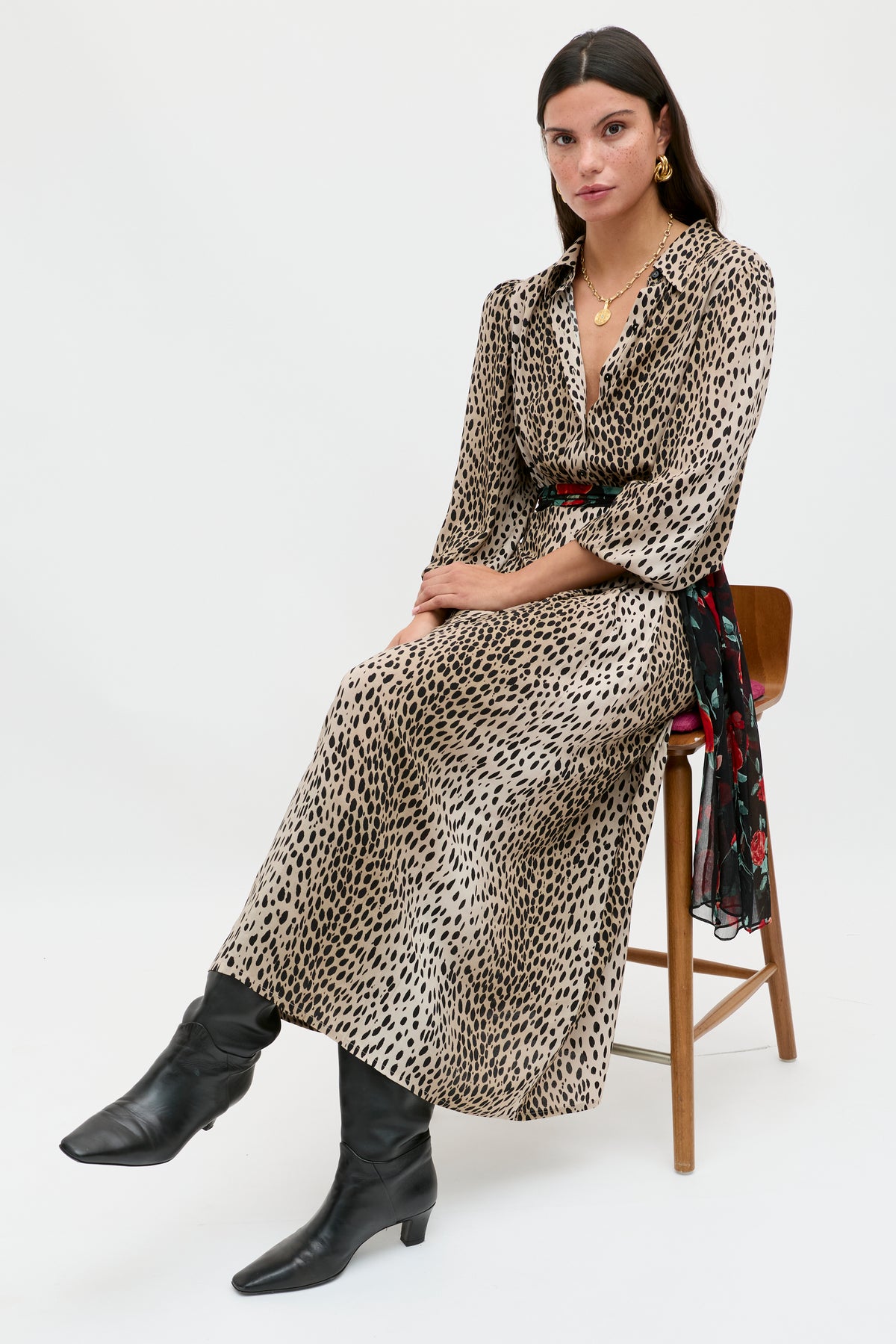 A woman sits on a wooden chair, wearing the Izzy - Antelope Spot Black shirt dress with black heeled boots, gold jewelry, and a scarf at her waist. The look is perfect for work or weekends. The background is plain white.