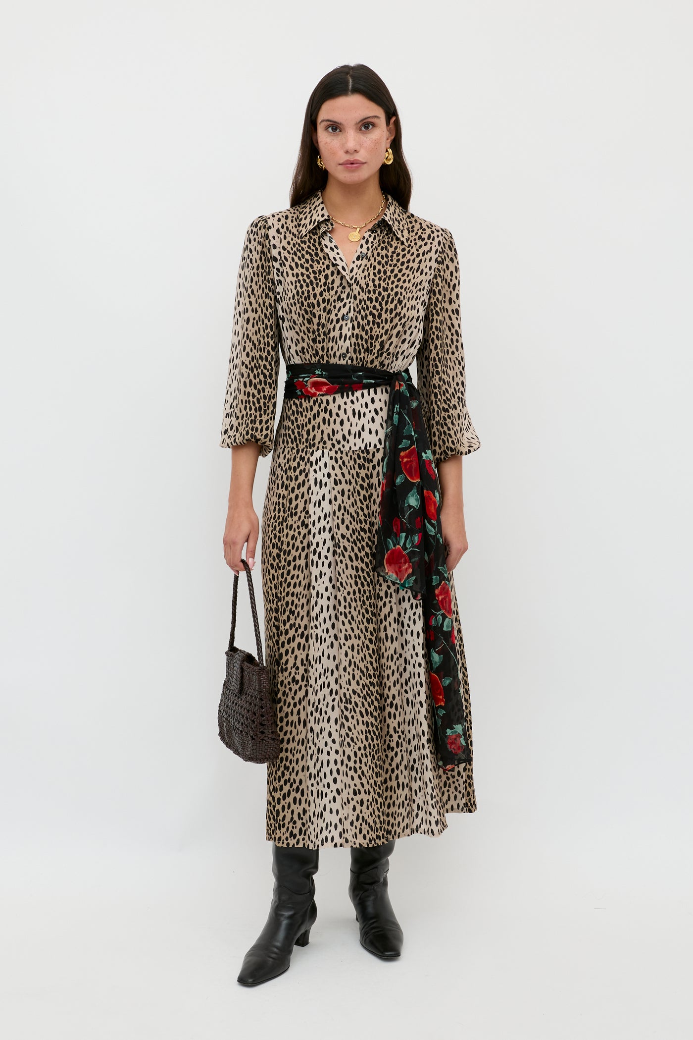 A woman poses against a white background wearing the Izzy - Antelope Spot Black long shirt dress with a floral scarf belt, black knee-high boots, gold jewelry, and a dark woven handbag—ideal for work or weekend style.