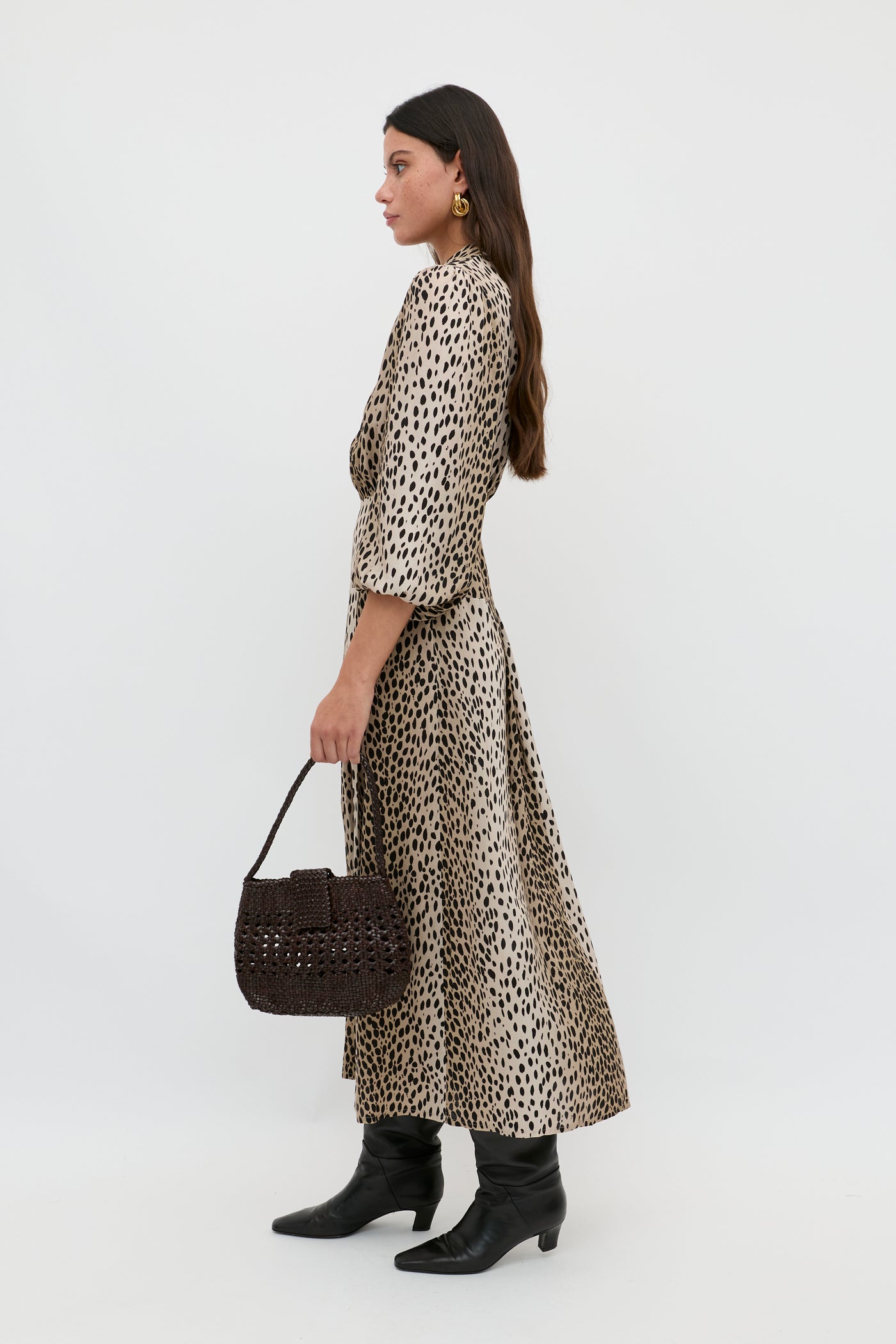 A woman wears the Izzy - Antelope Spot Black, a beige ankle-length shirt dress with black spots. Styled with black boots, gold earrings, and a dark woven handbag, she stands sideways against a plain white background.
