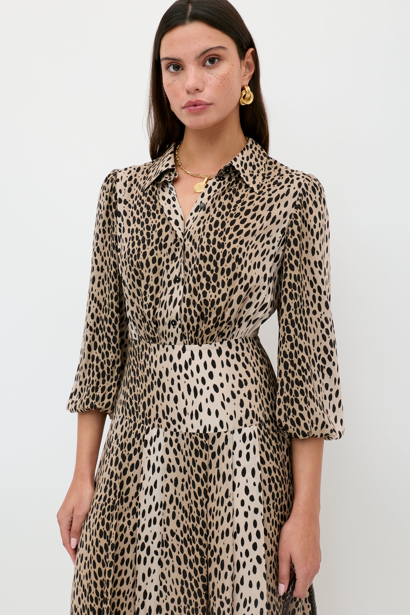 A woman with long brown hair wears the Izzy - Antelope Spot Black shirt dress, featuring a collared neckline and three-quarter sleeves—ideal for work or weekend. She poses against a white background, accessorized with gold earrings and a necklace.