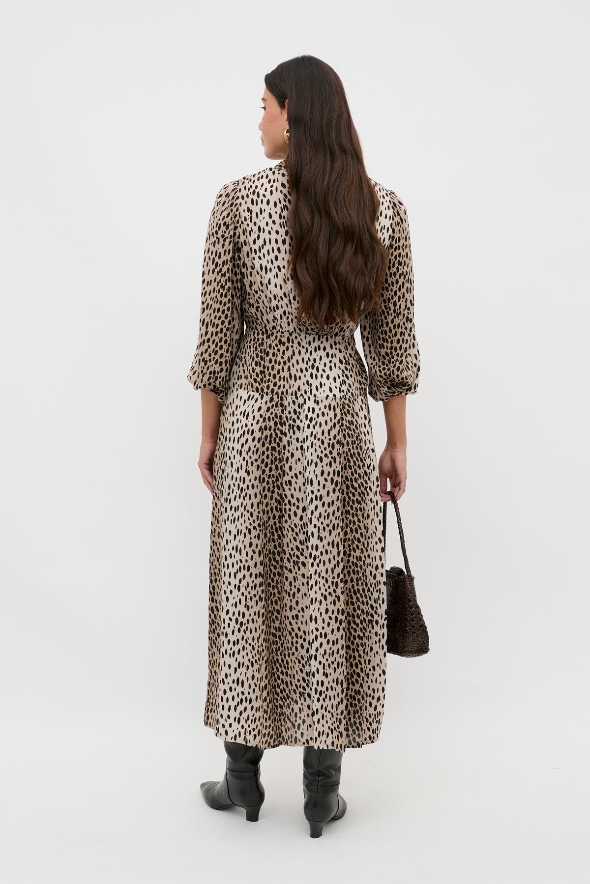A woman with long, wavy brown hair stands facing away in the Izzy - Antelope Spot Black shirt dress, paired with black boots and a textured dark handbag. Her effortless look works for both weekdays and weekends against a plain white background.